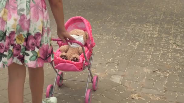 Een kind op straat met zijn favoriete speeltje met een beschermend masker. gezond kinderconcept. Meisje loopt in het park met een kinderwagen en een teddybeer met medisch masker. kind speelt een epidemie en beschermt speelgoed. — Stockvideo