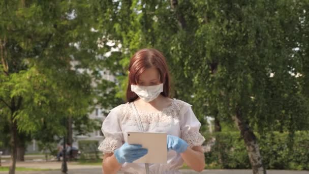 Zdravé děvče chodí v ochranné masce a lékařských rukavicích pracuje s tabletem v městském parku. COVID-19. nebezpečí viru. mladá podnikatelka pracující na dálku na ulici. Bezpečná a společenská vzdálenost. — Stock video