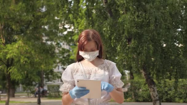 Una niña sana está caminando en máscara protectora y guantes médicos que trabajan con la tableta en el parque de la ciudad. COVID-19. peligro de virus. joven mujer de negocios trabajando remotamente en la calle. Distancia segura y social . — Vídeos de Stock