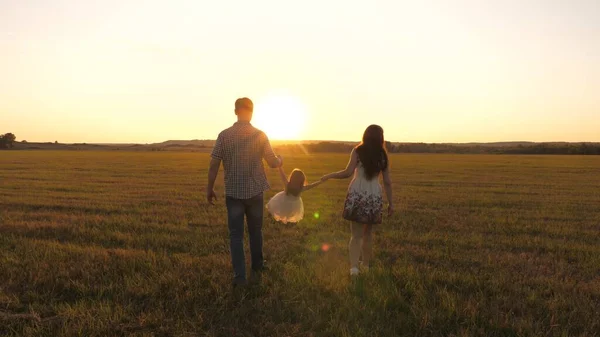 Petite fille joue et saute dans le champ tenant la main de maman et papa. en bonne santé Famille heureuse avec un enfant marche dans le parc au coucher du soleil. bébé et parent marchent au soleil. concept de famille heureuse et d'enfance — Photo