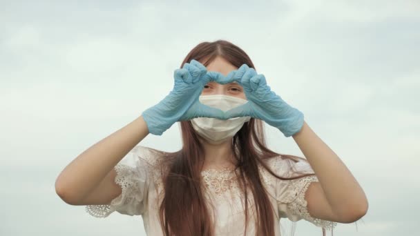 A jovem adora médicos. pandemia, covid 19. menina em uma máscara protetora e luvas médicas mostra símbolo do coração com as mãos. médico feminino mostrando dedos de coração e amor para pacientes . — Vídeo de Stock