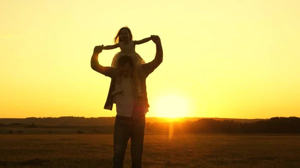 Papà porta sulle spalle del suo amato bambino, in raggi di sole. Padre cammina con la figlia sulle spalle in raggi di tramonto. bambino con genitori cammina al tramonto. famiglia felice che riposa nel parco. — Foto Stock