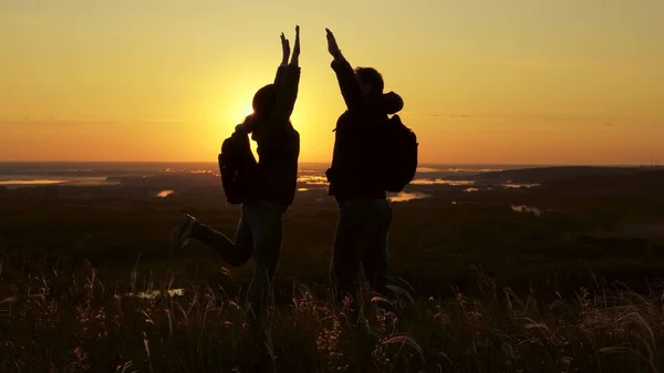 Travelers, a man and woman with backpack, go to edge of hill in rays of dawn, raise their hands, clap their hands and enjoy victory and beautiful sun and landscape. family travel and tourism concept — Stock Photo, Image