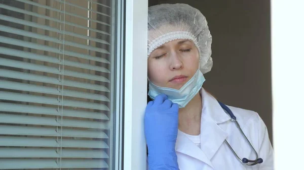 Femme fatiguée médecin en combinaison de protection regarde par la fenêtre, enlève son masque médical, ferme les yeux de la fatigue, respire l'air frais. Coronavirus pandémique. Dur journée de travail du médecin à l'hôpital. — Photo