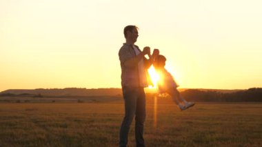 Sağlıklı aile ve çocukluk kavramı. Mutlu çocuk gün batımında babasıyla sahada sinek oyunu oynuyor. Baba, sevgili kızının kollarında uçarken gülümseyen sağlıklı çocuğunun etrafında dönüyor. Baba ve bebek yaz parkında.
