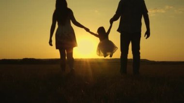küçük kızı güneşin arka planda parkta baba ve anne el ele atlayarak. Aile konsepti. çocuk gün batımı ışığında sahada baba ve anne ile oynar. Doğada küçük bir çocukla yürümek.