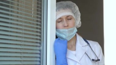Koruyucu takım elbiseli yorgun kadın camdan dışarı bakar, tıbbi maskesini çıkarır, yorgunluktan gözlerini kapatır, temiz hava soluyarak nefes alır. Salgın Koronavirüs. Hastanede çok çalışan bir doktor var..