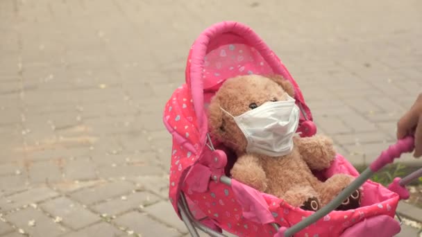 Kind spielt Epidemie und schützt Spielzeug. Kind auf der Straße mit seinem Lieblingsspielzeug in Schutzmaske. Konzept einer gesunden Kindheit. Kleines Mädchen spaziert mit Kinderwagen und Teddybär in medizinischer Maske durch Park. — Stockvideo