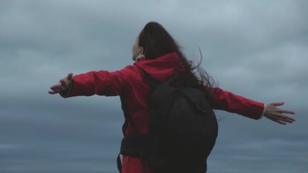 Glad flicka resenär går till kanten av berget med utsträckta armar, vinden viftar hennes hår. Gratis kvinna reser med en ryggsäck, ser från toppen av berget, njuter av landskapet och floden. — Stockvideo