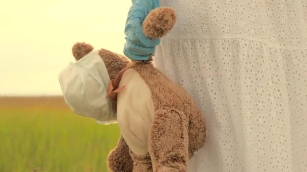 Девушка носит в руке игрушечного плюшевого мишку в медицинской маске в летнем парке. ребенок держит своего любимого плюшевого мишку в защитной маске во время карантина. Пандемический коронавирус Ковид-19. концепция детского здоровья — стоковое видео