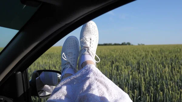 Legs of a girl in car window, riding car on country road past wheat field. free woman travels by car. healthy Young woman enjoys traveling by car, protruding her legs from an open window. travel