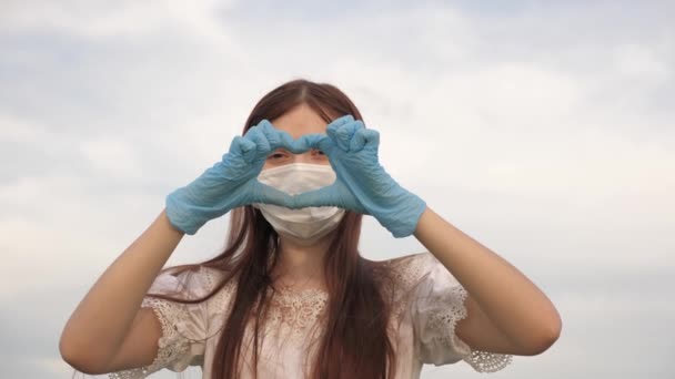 保護マスクと医療用手袋の女の子は手で心臓のシンボルを示しています。若い女性は医者が好きだ。パンデミック、 covid 19患者のための心と愛の指を示す女性医師. — ストック動画