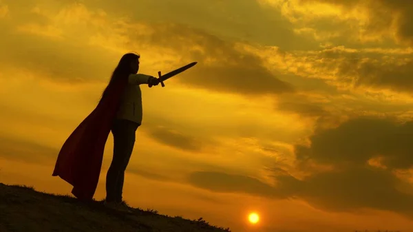Дівчина грає римського Леніна в яскравих променях сонця проти неба. вільна жінка лицар. супер жінка з мечем у руці і червоним плащем стоїть на горі під час заходу сонця. вільна жінка грає супергероя . — стокове фото