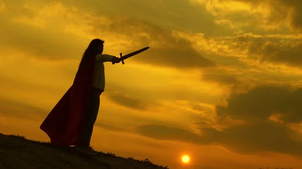Dziewczyna gra rzymskiego lenina w jasnych promieni słońca przeciwko niebu. wolny rycerz. super kobieta z mieczem w ręku i w czerwonym płaszczu stoi na górze w świetle zachodu słońca. wolna kobieta gra superbohatera. — Zdjęcie stockowe