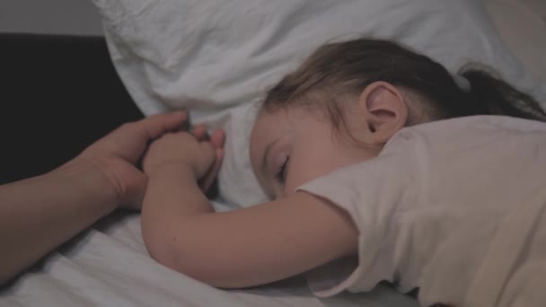 Pequena filha dorme na cama com uma mãe carinhosa ela segura a mão de seu filho. mãe e bebê estão descansando na cama. família feliz dormindo de manhã no sofá — Vídeo de Stock