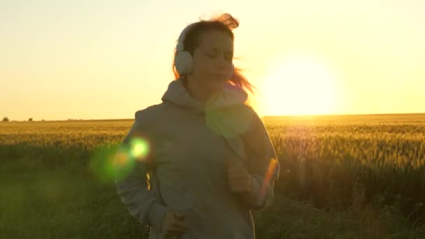 Belle fille en bonne santé faisant du jogging de remise en forme sur un champ de blé. Entraînement. Jeune femme libre court en été à l'aube écoute de la musique avec des écouteurs. Jogger fille respire l'air frais — Video