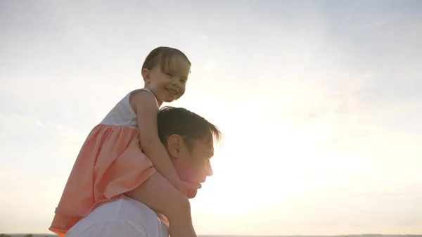 Criança com pais anda ao pôr-do-sol. Papai carrega nos ombros de seu filho amado, em raios de sol. O pai caminha com a filha sobre os ombros em raios de sol. família feliz descansando no parque . — Fotografia de Stock