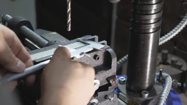 Mecánico mide el detalle de la pinza en el taller. Trabajador en una fábrica en un taller de cerrajería. Taladradora, taladradora para empleados en una placa de acero plana con un taladro de mesa . — Vídeo de stock