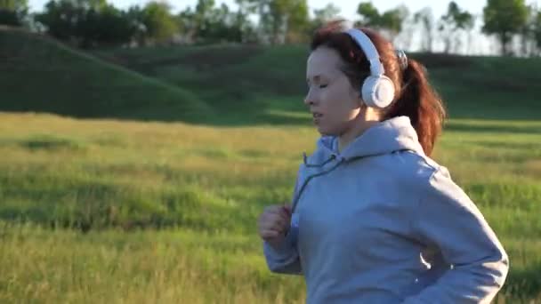 Νεαρή γυναίκα εκπαιδεύεται το καλοκαίρι στο πάρκο την αυγή σε ακουστικά με μουσική. Δωρεάν όμορφο κορίτσι ασχολείται με τη φυσική κατάσταση. Το κορίτσι που κάνει τζόγκινγκ αναπνέει καθαρό αέρα. αθλήματα για καύση θερμίδων και απώλεια βάρους — Αρχείο Βίντεο