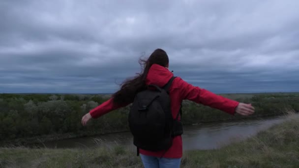 Mujer joven viaja con una mochila, mira desde la cima de la montaña, disfrutando del paisaje y el río. libre chica feliz viajero va a la orilla de la montaña con los brazos extendidos, el viento está agitando su pelo. — Vídeos de Stock