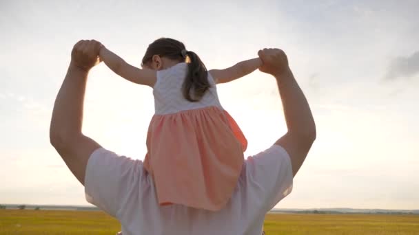 Padre camina con su hija sobre sus hombros en rayos de atardecer. Papá lleva sobre los hombros de su amado hijo, en rayos de sol. niño con padres camina al atardecer. familia feliz descansando en el parque. — Vídeo de stock