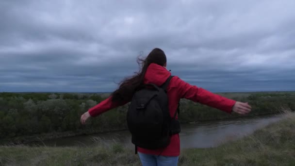 Mujer joven viaja con una mochila, mira desde la cima de la montaña, disfrutando del paisaje y el río. libre chica feliz viajero va a la orilla de la montaña con los brazos extendidos, el viento está agitando su pelo. — Vídeos de Stock