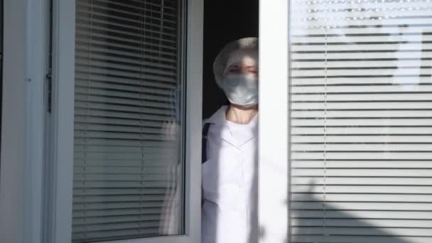 Zmęczona lekarka w kombinezonie ochronnym patrzy przez okno, zdejmuje maskę medyczną, zamyka oczy przed zmęczeniem, oddycha świeżym powietrzem. Ciężki dzień pracy lekarza w szpitalu. Pandemiczny koronawirus — Wideo stockowe