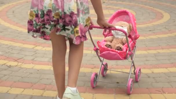 Kid on street with his favorite toy in a protective mask. healthy childhood concept. little girl walks in park with a pram and a teddy bear in medical mask. child plays an epidemic and protects toy. — Stock Video