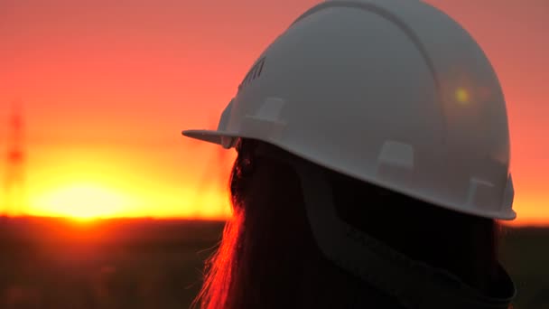 Een vrouwelijke energietechnicus in een witte helm inspecteert een hoogspanningslijn. Hoogspanningsleidingen bij zonsondergang. Distributie en levering van elektriciteit. schone energie — Stockvideo