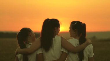 Anne ve kızları gün batımında tarlada, mutlu bir aile. Anne yetişkin çocukları kucaklıyor, onlar güzel gün batımını takdir ediyorlar. Mutlu aile ve çocuk kavramı. Anne ve kızları parkta, tarlada, güneşte yürüyorlar.