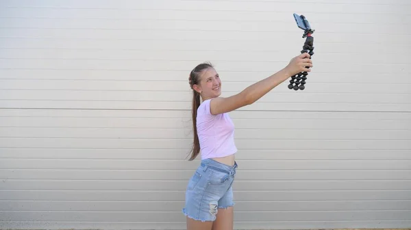 Hermosa chica hace selfie al aire libre. adolescente es fotografiado en un teléfono inteligente utilizando trípode pequeño. joven bloguera dirige una videoconferencia en línea desde la calle de la ciudad. tecnologías digitales modernas — Foto de Stock