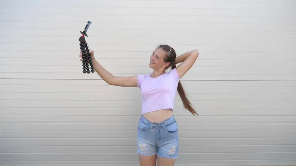 Hermosa chica hace selfie al aire libre. adolescente es fotografiado en un teléfono inteligente utilizando trípode pequeño. joven bloguera dirige una videoconferencia en línea desde la calle de la ciudad. tecnologías digitales modernas — Foto de Stock