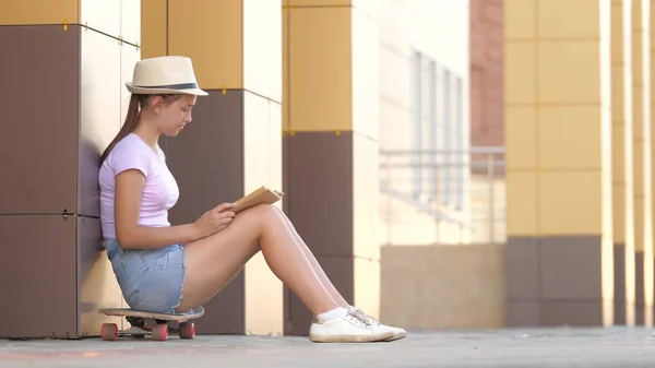 Una chica con sombrero lee un libro, en la ciudad cerca del edificio de la escuela sentado en un monopatín. Adolescente está estudiando una lección. colegiala con un libro de texto en sus manos. concepto de educación y desarrollo —  Fotos de Stock