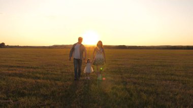 Küçük kız, anne ve babasını elleriyle tutuyor ve sarı güneş ışınlarıyla parktaki çayırlarda zıplıyor. Aile mutluluğu. Sağlıklı bebek gün batımında anne ve babayla sahada oynuyor. Doğada yürüyen çocuk.