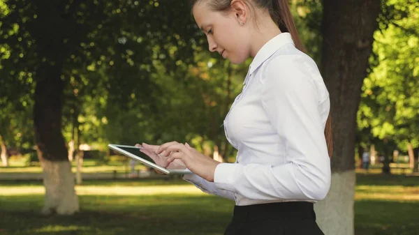 Portrait of a young girl with a tablet in her hand. female student teaches lessons on the street with a tablet in the park. Happy woman reading good news in an external message.