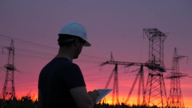 Beyaz kasklı erkek güç mühendisi güç hattını kontrol ediyor, elinde bir tablet tutuyor. Gün batımında yüksek voltajlı elektrik hatları. Dağıtım ve elektrik tedariği. temiz enerji