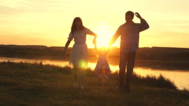 Küçük kız anne ve babasını ellerinden tuttu ve sarı güneşin altında kumsala atladı. Aile mutluluğu. Sahada gün batımında anne ve babayla oynayan sağlıklı bir bebek. Doğada yürüyen çocuk.