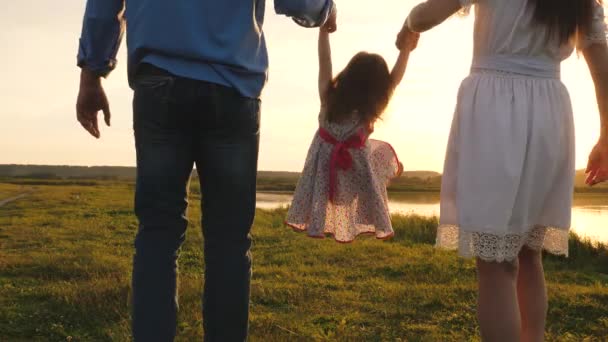 Zdravé dítě si hraje s tátou a mámou na hřišti při západu slunce. Dcera drží mámu a tátu za ruce a skočí na pláž pod žlutým sluncem. Chodící dítě v přírodě. šťastný, zdravý rodinný koncept. — Stock video