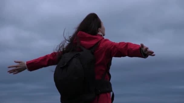 Свободная девушка путешественник идет к краю горы с протянутыми руками, ветер развевает ее волосы. Свободная женщина путешествует с рюкзаком, смотрит на облака, наслаждаясь красивым пейзажем. — стоковое видео