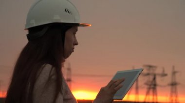Beyaz kasklı bir kadın güç mühendisi tabletteki elektrik sensörlerinden elde edilen verileri kullanarak güç hattını kontrol ediyor. Gün batımında yüksek voltajlı elektrik hatları. Dağıtım ve elektrik tedariği. temiz enerji