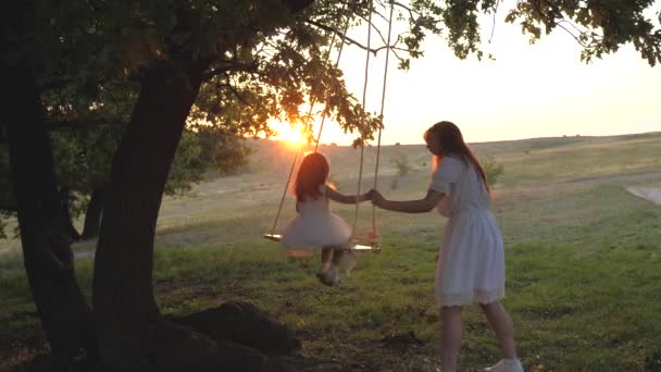 Mãe treme sua filhinha saudável em um balanço sob uma árvore ao sol. mãe brinca com seu filho, eles balançam na corda em um galho de carvalho na floresta. A família livre está se divertindo no parque. — Vídeo de Stock