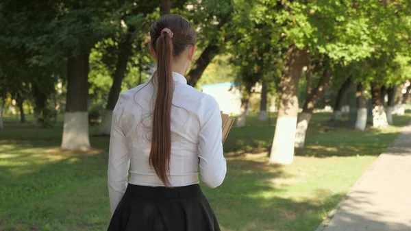 Una estudiante camina por la calle con libros de texto en sus manos en el parque de verano. colegiala en la ciudad. chica adolescente se apresura a la escuela con libros. —  Fotos de Stock