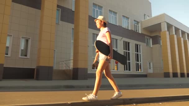 Sportos és egészséges életmód a modern tinédzser. Ingyenes tini lány megy egy gördeszkával a kezében az utcán a városban. boldog gördeszkás utazik az úton a szabadban a parkban. — Stock videók
