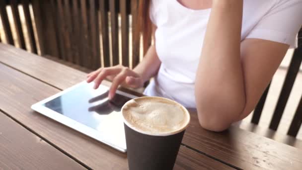 Una chica en un café de verano utiliza una tableta y bebe delicioso café aromático. mujer charlando, tomando café y relajándose. mujer joven en un café trabaja con un gadget. — Vídeo de stock