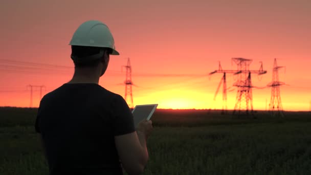 Électricien travaille sur la ligne électrique. un ingénieur en énergie dans un casque blanc vérifie la ligne électrique, tient la tablette dans ses mains. Lignes électriques à haute tension au coucher du soleil. Distribution et fourniture d'électricité. — Video