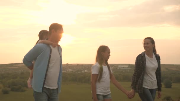 Gezond glimlachend gezin, hand in hand, lopend over het veld bij zonsondergang in de bergen, kleine dochter rijdt op papa 's rug. gelukkige kinderen en ouders wandelen in stralen van mooie zon, reizen op vakantie — Stockvideo