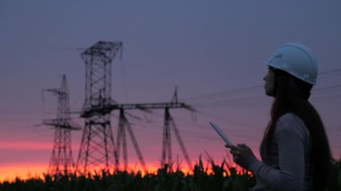 Beyaz kasklı bir kadın güç mühendisi tabletteki elektrik sensörlerinden elde edilen verileri kullanarak güç hattını kontrol ediyor. Gün batımında yüksek voltajlı elektrik hatları. Dağıtım ve elektrik tedariği. temiz enerji