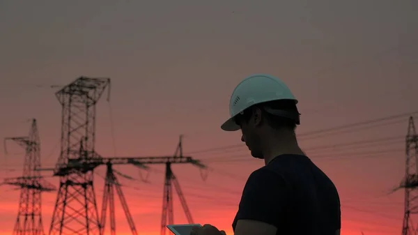 O engenheiro de energia num capacete branco verifica a linha eléctrica, segura um tablet nas mãos. Linhas eléctricas de alta tensão ao pôr-do-sol. Distribuição e fornecimento de electricidade. energia limpa — Fotografia de Stock