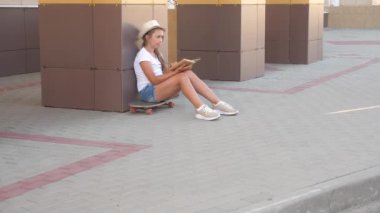 Elinde ders kitabı olan bir kız öğrenci. Şapkalı bir kız kitap okuyor, okul binasının yakınındaki kaykayın üzerinde oturuyor. Genç bir ders çalışıyor. eğitim ve geliştirme kavramı