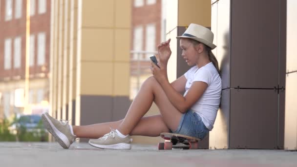 Modieus meisje leest met behulp van moderne technologie smartphones. Een jong duizendjarig meisje zit op een skateboard en kijkt naar een tablet. Tienersport en educatieve levensstijl. concept van online onderwijs — Stockvideo